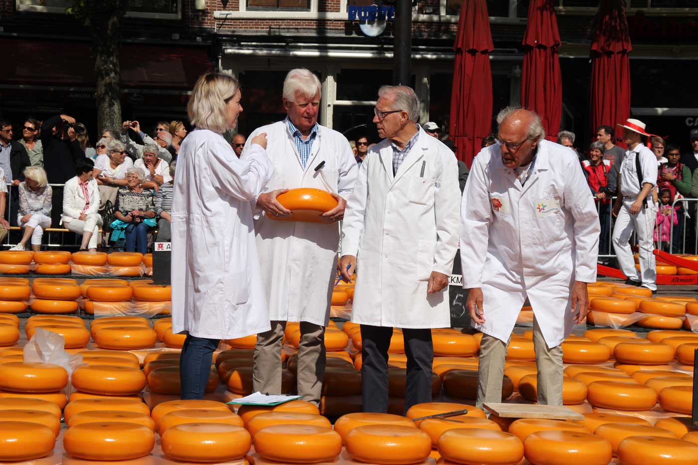 По центру площади с сырами стоят 4 эксперта по сыру в белых халатах и осматривают одну из головок.