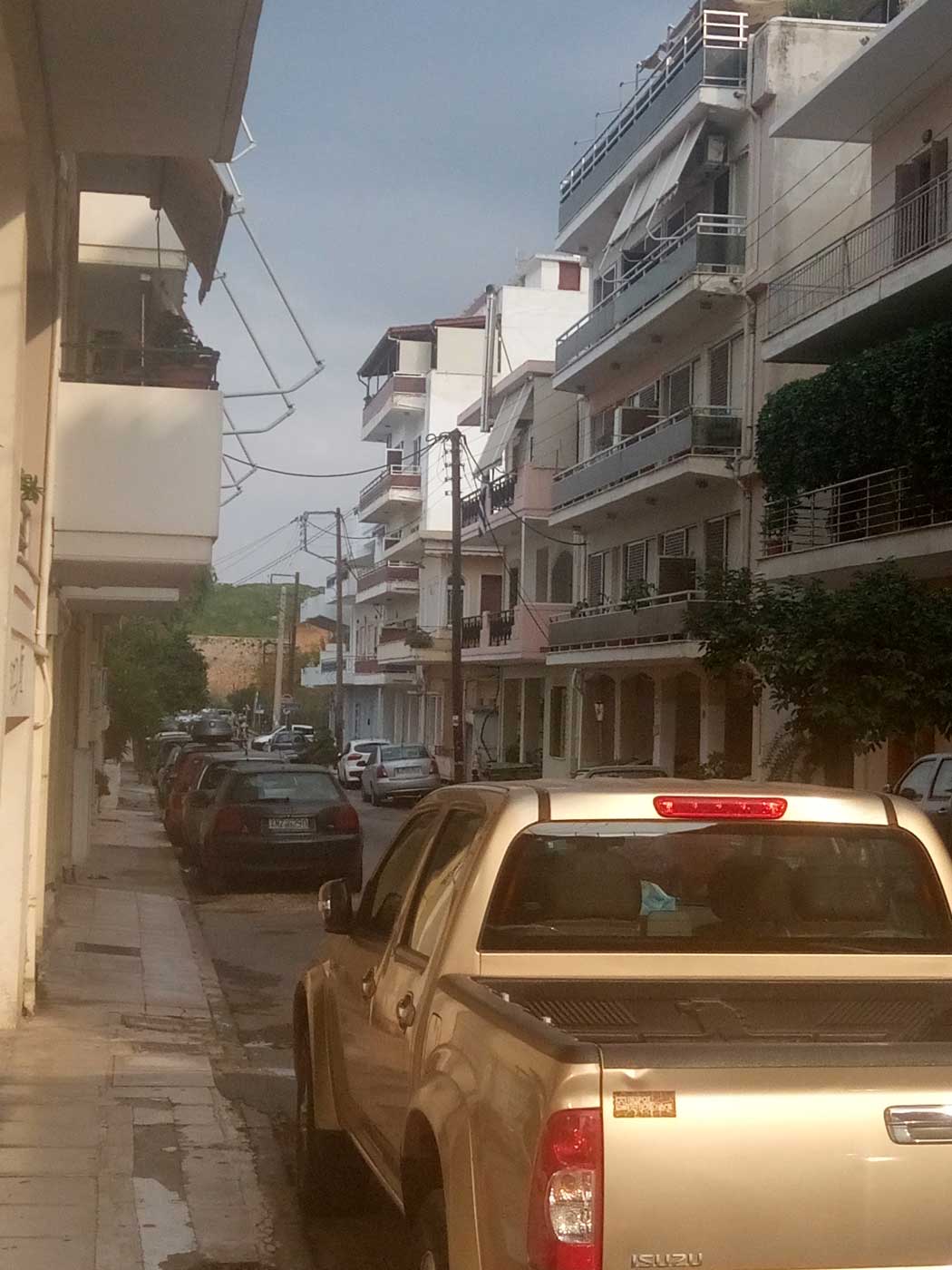 Узкая улица, вдоль которой стоят невысокие белые дома. По обеим сторонам плотно припаркованы автомобили.